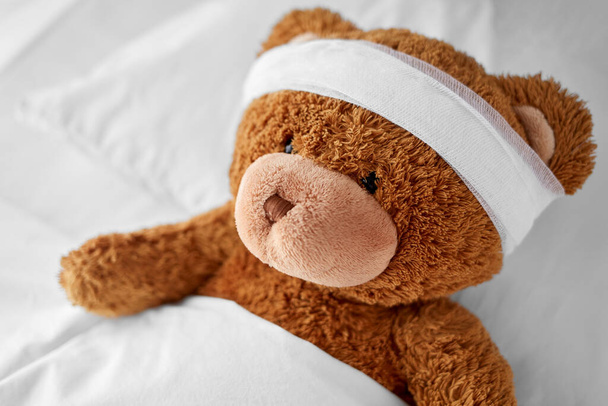 ill teddy bear toy with bandaged head lying in bed - Фото, изображение