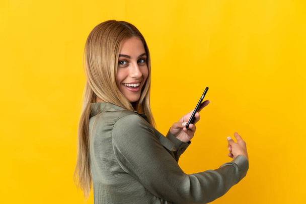 Молода блондинка ізольована на жовтому тлі, використовуючи мобільний телефон і вказуючи назад
 - Фото, зображення