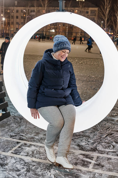冬のスポーツウェアとアイススケートの笑顔のシニア女性の完全な長さの肖像画は、冬の夜に市のスケートリンクでリングベンチに座っています。アクティブ・エイジング、ボディ・ポジティブ・コンセプト - 写真・画像