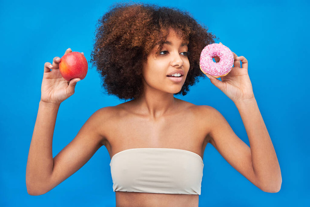 Кудрявая женщина позирует с яблоком и пончиком у голубой стены - Фото, изображение