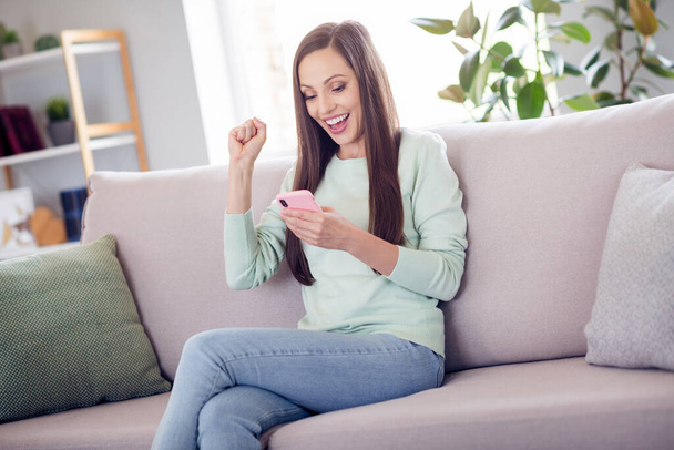 Photo of hooray millennial brunette lady sit on sofa with telephone wear blue shirt jeans at home alone - Fotoğraf, Görsel