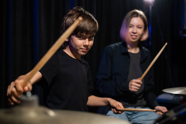 Молодая кавказка учит мальчика играть на барабанах в студии на черном фоне. Студентка музыкальной школы - Фото, изображение
