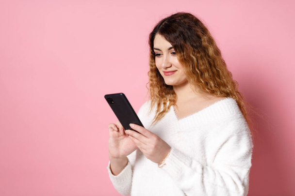 A cheerful charming young woman, standing isolated on a pink background with a smartphone in her hands, looks into the camera and smiles. - Foto, Imagen