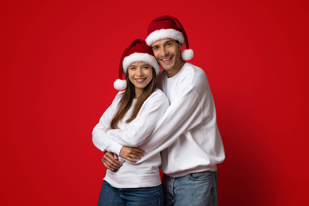 Retrato de abraçar jovem casal em chapéus de Santa posando sobre fundo vermelho - Foto, Imagem