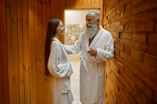 Hombre mayor y mujer joven en albornoz hablando de pie en el salón de sauna spa compartiendo impresiones - Foto, imagen