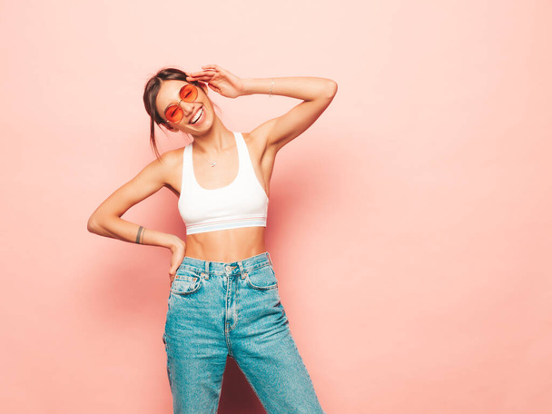 Beautiful smiling woman dressed in white jersey top shirt and jeans. Sexy carefree cheerful model having fun indoors.Adorable and positive female posing near pink wall in studio. In sunglasses - Φωτογραφία, εικόνα