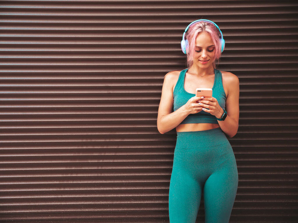 Fitness smiling woman in green sports clothing with pink hair. Young beautiful model with perfect body.Female in the street near roller shutter wall.Listening music in wireless headphones - Foto, Imagen