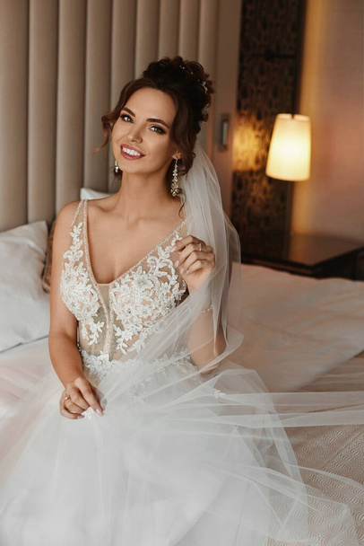 Adorabile giovane sposa con acconciatura elegante indossa abito alla moda e orecchini seduti sul letto - Foto, immagini
