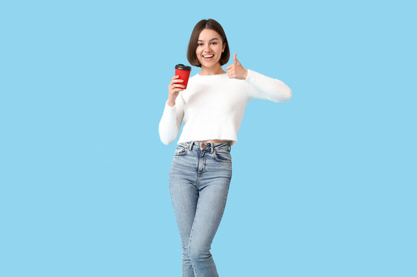 Mooie vrouw met kopje koffie tonen duim-up op kleur achtergrond - Foto, afbeelding