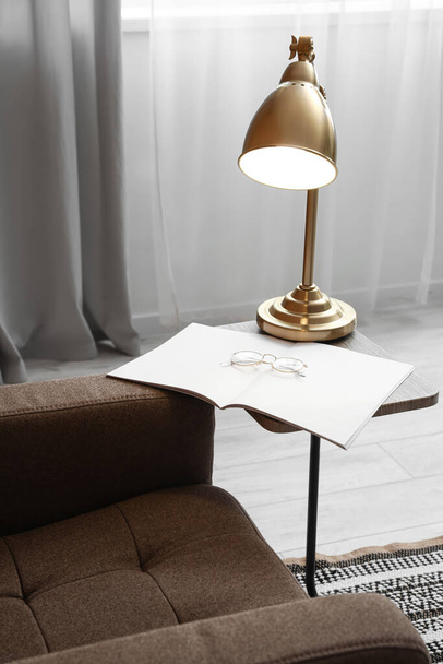 Gloeilamp, boek en bril op salontafel bij fauteuil in woonkamer - Foto, afbeelding