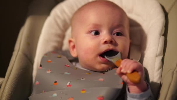Kleine jongen die op een voedselstoel zit te knagen aan een rubberen lepel. Schattige baby kinderziektes en bijtende lepel na het eten. - Video
