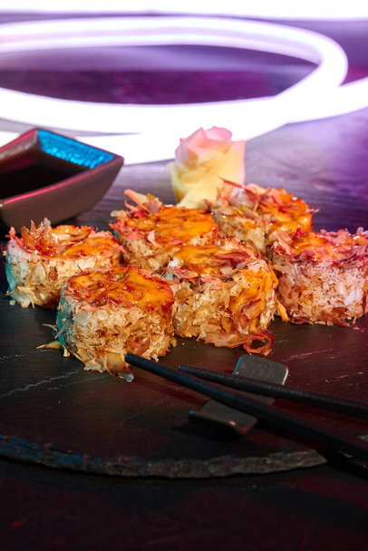 Detailní záběr uramaki rolí s roztaveným sýrem a bonitovými vločkami na břidlicové desce s jasným LED páskem - Fotografie, Obrázek