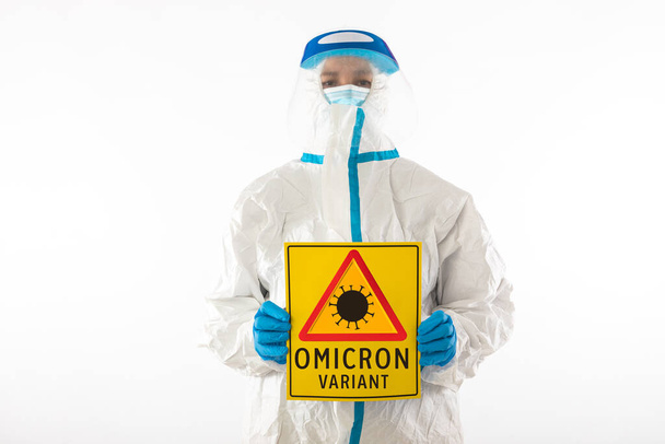 Medical nurse in personal protective equipment and latex gloves, holding a yellow sign with a hazard symbol that reads: 'VARIANT OMICRON'. Coronavirus, pandemic and health concept. - Foto, Imagen