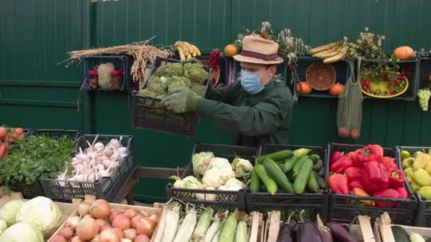 Broccoli al mercato agricolo. Un venditore maschio mette una scatola di broccoli sul banco.La fotocamera si muove senza intoppi lungo le scatole con una varietà di verdure.Il venditore in una maschera medica protettiva.. - Filmati, video