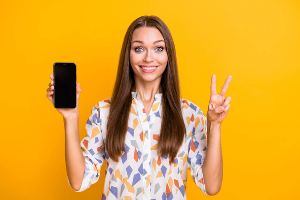 Photo of young girl positive smile show v-sign cool hold cellphone mobile isolated over yellow color background - Zdjęcie, obraz