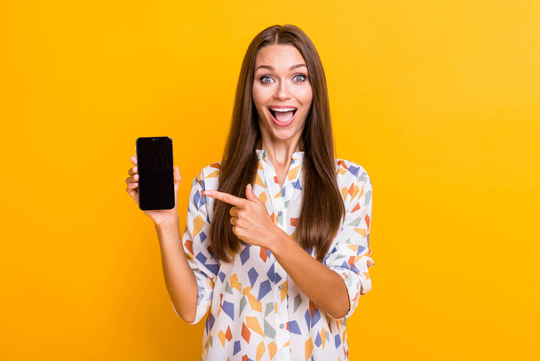 Photo portrait of excited girl pointing finger at phone with blank space holding in one hand isolated on vivid yellow colored background - Photo, image