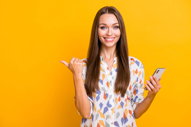 Photo portrait of girl pointing thumb at blank space holding phone in one hand isolated on vivid yellow colored background - Fotoğraf, Görsel