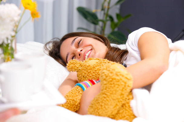 Young pretty happy woman lying in her bed and sleeping holding her favorite soft toy bear, dreaming something beautiful. - Foto, Bild