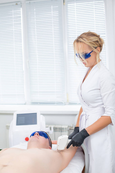 A pretty cosmetologist and her client in blue safety glasses during the procedure of laser hair removal from the armpit of a young man - Fotoğraf, Görsel