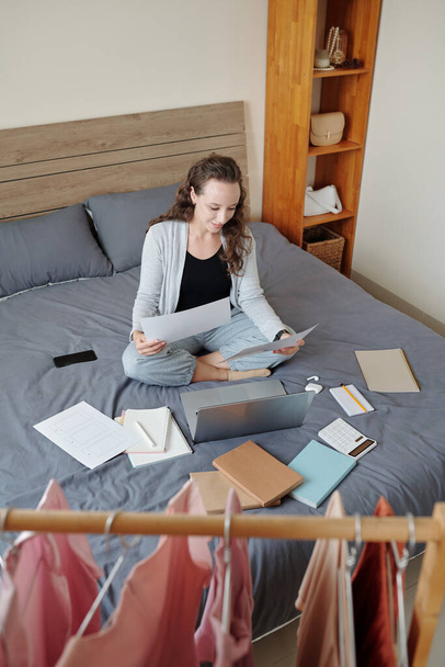 Mujer de negocios positiva sentada en la cama en casa y trabajando con documentos, vista desde arriba - Foto, Imagen