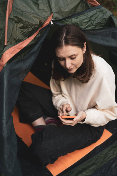 A young tourist sits in a tent in nature and looks at the phone. Surfing the Internet outside the city on hike - Фото, изображение