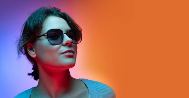 Studio shot of beautiful young girl waering sunglasses isolated on gradient blue-orange color background in neon. - Foto, Imagem