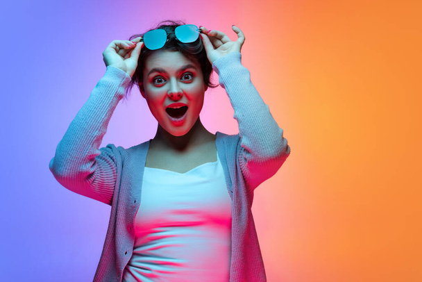 Studio shot of beautiful young girl waering sunglasses isolated on gradient blue-orange color background in neon. - Φωτογραφία, εικόνα