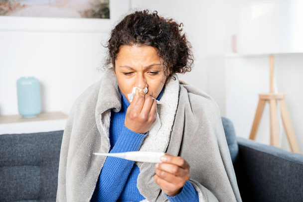 Черная женщина страдает от простуды и гриппа дома, проверяя термометр - Фото, изображение