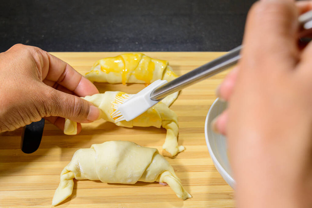 A high angle shot of a chef painting the ham and cheese croissants with egg yolk on a wooden board - Foto, Imagem