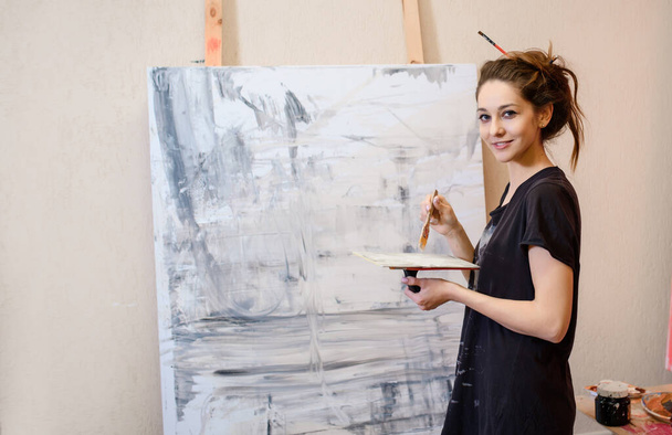 Siyah tişörtlü genç güzel ressam fırçasıyla soyut bir resim çiziyor ve stüdyosundaki kameraya bakıyor. - Fotoğraf, Görsel