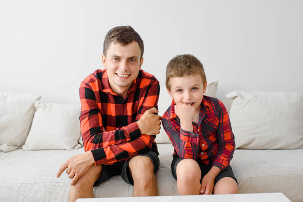 Mutlu genç baba ve oğlu, aynı ekose gömleklerle kanepede oturuyor ve kameraya bakıyorlar. Mutlu aile kavramı - Fotoğraf, Görsel