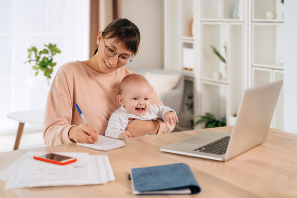 Vidám üzleti anya fogja a babáját, miközben távolról dolgozik az otthoni irodájában. Boldog egyedülálló anya mosolyog, jegyzetel, van online osztály. Fiatal női szabadúszó kiegyensúlyozó munka és anyaság - Fotó, kép