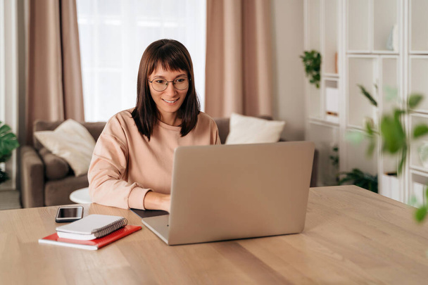 Kellemes boldog fiatal nő szabadúszó dolgozik laptop otthon. Vonzó üzletasszony szemüvegben online tanul, laptopszoftverrel, webes szörfözési információval vagy online vásárlással - Fotó, kép