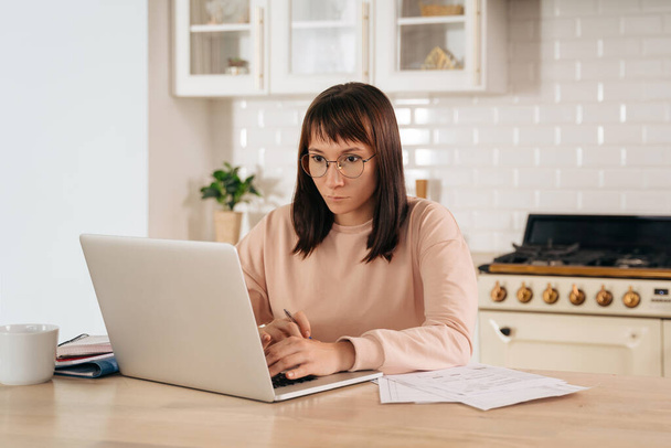 Serious woman wearing glasses working on laptop online, sitting at table in kitchen, looking at computer screen. Focused female using internet for distance education, searching information, up-skill - Photo, Image