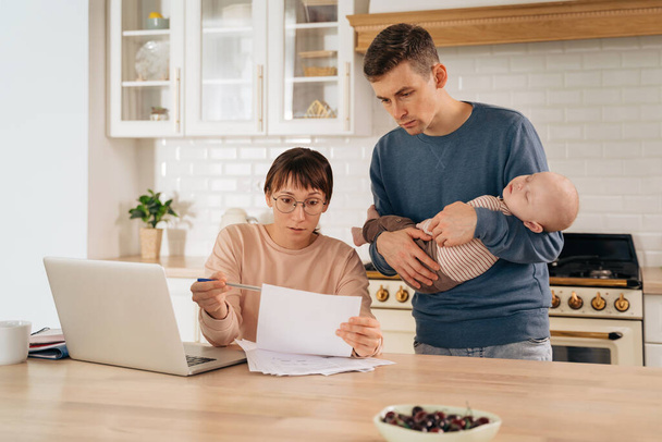 Business and family. Millennial parents with sleeping infant baby son working on laptop in kitchen, thinking of plans or financial problems, paying domestic bills, discussing family tasks and deals - Foto, Imagem