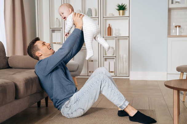 Happy young father is playing with his newborn baby boy on floor in living room at home. Cheerful dad holding his cute laughing toddler in his arms. Single father, fatherhood, paternity leave concept - Photo, Image