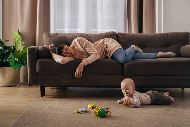 Młode zmęczone matki cierpiące na brak snu, śpiące na kanapie, podczas gdy jej małe niemowlę bawi się na podłodze. Wyczerpana mama przeżywa depresję poporodową, nie chce bawić się z synem - Zdjęcie, obraz