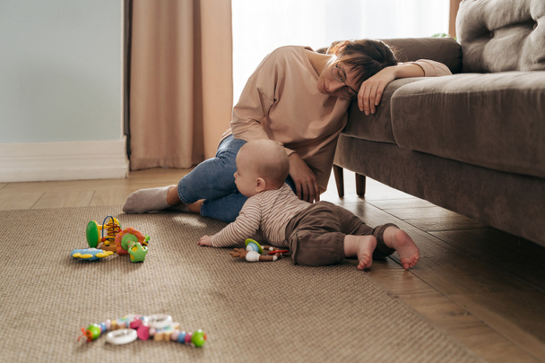 Young exhausted mother is very tired, fell asleep on sofa near her playing infant baby, suffering from postpartum depression, difficulties of motherhood, sleepless nights - Photo, Image
