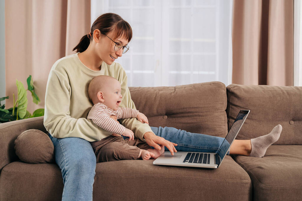 Happy young woman working from home with her adorable cheerful baby, sitting on couch, using laptop, having a video conference call. Busy multitasking mom working remotely with infant son. Motherhood - Foto, Bild