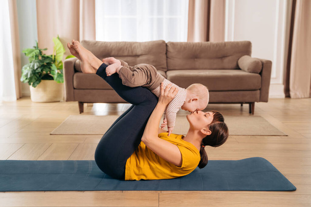 Sport with baby. Fit mom kissing her toddler son while holding him on her legs and doing postpartum recovery exercise on fitness mat at home - Photo, Image