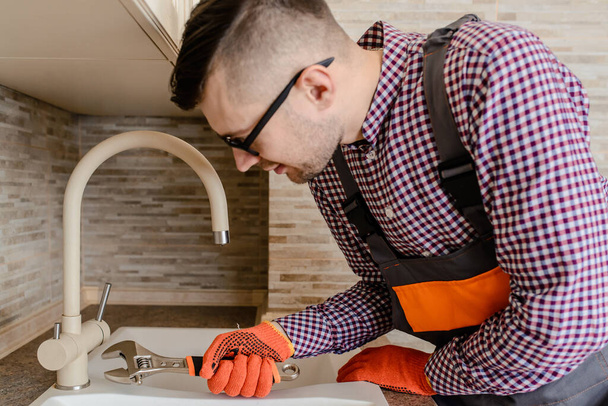 Young man plumber installer fixes with wrench the water tap pipe in vintage kitchen - Photo, Image