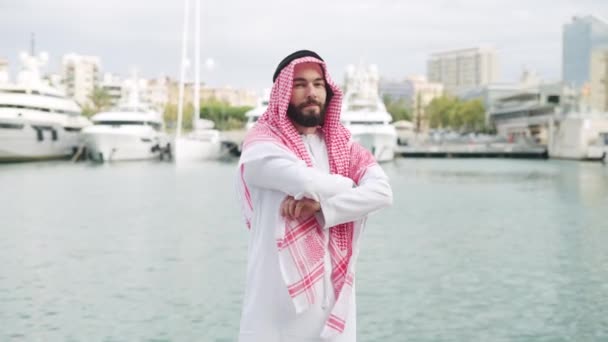 Szczęśliwy uśmiechnięty saudi mężczyzna pozowanie na zewnątrz w tradycyjnym stroju z ramionami skrzyżowane w porcie - Materiał filmowy, wideo