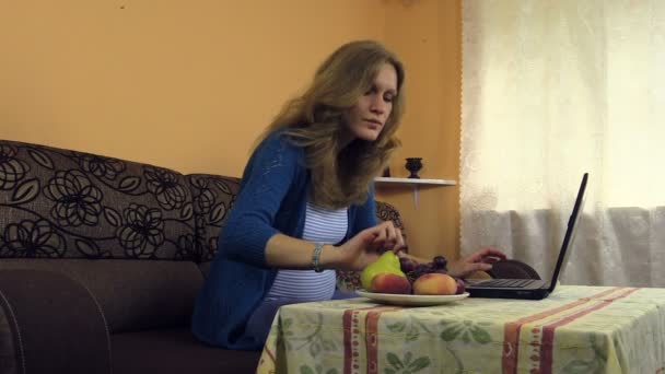 妊娠中の女性を扱う自宅でラップトップ、スナック ブドウ、フルーツの花瓶 - 映像、動画