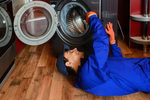 Mladý muž instalatér údržbář ležící na dřevěné podlaze v modrých montérkách opravit / opravit pračku v kuchyni. - Fotografie, Obrázek