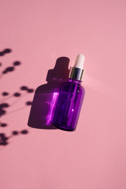 Purple glass dropper bottle with metallic lid. Top view on pink background with plant shadow. Skincare cosmetic. Beauty concept for face body care - Foto, afbeelding