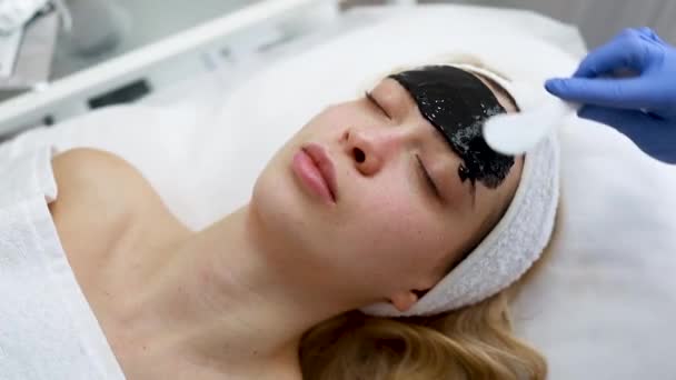 Ο Beautician απλώνει στο πρόσωπο του ασθενούς μια μαύρη μάσκα στο δέρμα με μια σπάτουλα. Διαδικασία αναζωογόνησης και καθαρισμού προσώπου - Πλάνα, βίντεο
