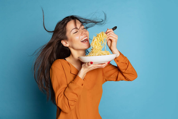 Portrait of beautiful young girl eating delicious Italian pasta isolated on blue studio background. World pasta day - 写真・画像