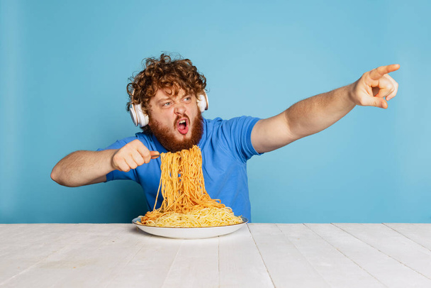 Excited young hairy red-bearded man tasting large portion of noodles, pasta isolated on blue studio background. - Foto, Imagen