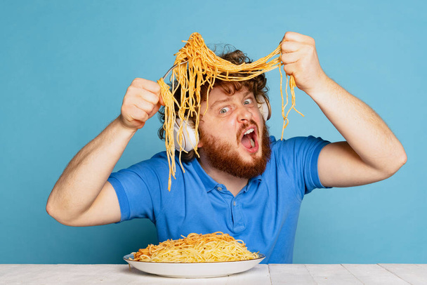 Crazy young hairy red-bearded man tasting large portion of noodles, pasta isolated on blue studio background. - Fotografie, Obrázek