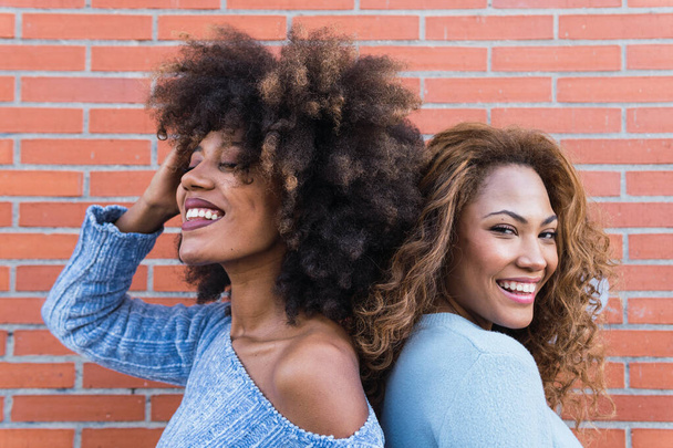 Vista laterale di donne africane americane sorridenti con lunghi capelli afro, trucco glamour e camicia blu in posa nel muro di mattoni. Ritratto di bellezza di ragazze naturali africane in piedi schiena a schiena - Foto, immagini